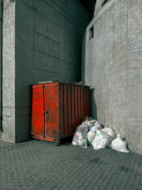 The image shows a red metal rubbish skip with a weathered surface, positioned against a dark green wall on the left side and a grey concrete wall on the right. The skip is partially filled with litter and debris. In front of the skip, there are several clearly visible white plastic bags containing waste, some slightly misshapen and bulging, indicating they are filled with rubbish. The ground is paved with dark grey interlocking bricks, and the area appears to be an alley or side passage, with a small rectangular window above the skip on the grey wall. The scene is lit evenly, suggesting natural daylight. This image depicts a typical private waste storage situation, often used as an alternative to regular rubbish collection services, supported by companies like Rubbish Clearance Putney, for on-site or independent rubbish disposal, aligning with the context of avoiding hidden fees in Putney rubbish clearance.
