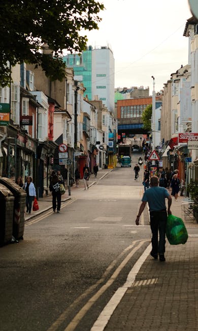 A busy urban street scene in Putney with a mix of pedestrian activity and commercial buildings. In the foreground, a man wearing a teal shirt and dark trousers is walking away from the camera, carrying a large green reusable shopping bag. To the left, pedestrians including a person with a backpack and individuals walking in groups are visible along the sidewalk. The street is flanked by a variety of small shops and cafes with storefronts, awnings, and signage, some indicating food and retail businesses. The buildings are a mixture of older, traditional brick structures and modern glass-fronted offices, with a noticeable large turquoise and white office building in the background. The street includes pavement, double yellow lines, and a slight incline leading towards a larger building complex at the top of the slope. Overhanging trees cast partial shadows, and there are several parked cars along the roadside. The scene is illuminated by natural daylight, suggesting a partly cloudy day, with a quiet atmosphere suitable for a private rubbish clearance or alternative waste handling in a typical residential and commercial area. Rubbish Clearance Putney often manages waste removal for local properties in similar urban environments, reflected in the clean, organized surroundings.
