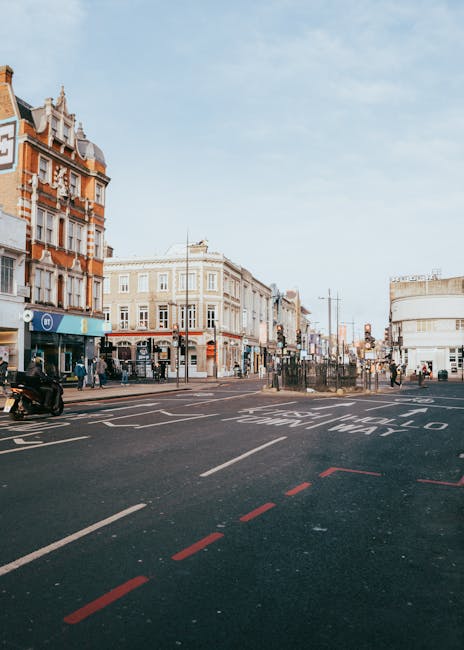 A busy street scene on Putney High Street shows a wide asphalt road with clear white and red lane markings, including a dashed red line indicating a no-parking zone or restricted area. In the foreground, the road is smooth and dark with visible traffic lines, leading towards a cluster of mixed-use buildings in the background. These structures feature detailed masonry, brickwork, and large glass windows; one building on the left exhibits intricate decorative elements and a prominent cornice with a pointed top. Pedestrians are seen walking along the sidewalks, some near shop entrances, while others cross the street at designated crossing points. To the left, a motorbike is parked close to the curb, and a few street furniture pieces, such as lampposts and traffic signals, are present. The sky overhead is partly cloudy with a soft, natural light illuminating the scene, emphasizing the urban environment suitable for various forms of private waste collection or rubbish clearance services, as might be offered by Rubbish Clearance Putney in managing street-level waste and debris within this commercial and residential area.