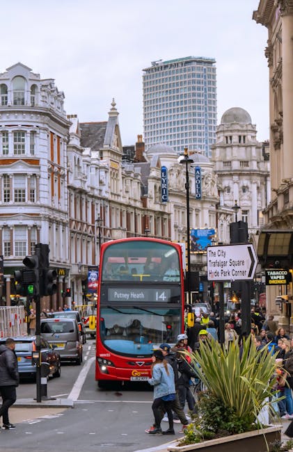 A busy urban street scene in Putney featuring a double-decker bus with a red exterior and a large front windshield displaying route information to Putney Heath. The bus is positioned centrally, surrounded by a variety of vehicles including cars and taxis, as well as numerous pedestrians walking along the sidewalks. The foreground includes a landscaped area with spiky, green foliage plants, and a traffic light on the left side. The background showcases historic white and beige Victorian-style buildings with ornate architectural details, bay windows, and decorative elements, alongside a modern high-rise office tower with a glass and steel facade. Street signs and lampposts are visible, contributing to the street's busy atmosphere. The scene is illuminated by natural daylight with an overcast sky, creating a neutral, professional tone suitable for an article on alternative rubbish removal services or on-site clearance in Putney, complemented by the company's presence in the area.