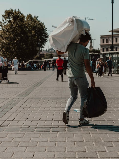 A young man is walking on a paved urban square carrying a large white plastic bag over his right shoulder and a smaller black bag in his left hand. He is dressed in casual attire, including a grey T-shirt, light grey jeans, and sneakers. The square is busy with pedestrians, some standing and others walking, and is surrounded by trees and historic-style buildings. Parked cars are visible along the street in the background. The scene is well-lit with natural daylight, and the atmosphere suggests a typical day of outdoor activity. This image reflects scenarios where individuals may be involved in personal waste disposal or recycling, aligning with private rubbish collection or alternative waste handling services such as those offered by Rubbish Clearance Putney. The visual emphasizes the movement and effort associated with rubbish removal outside of municipal collection, demonstrating practical, on-site clearance efforts.